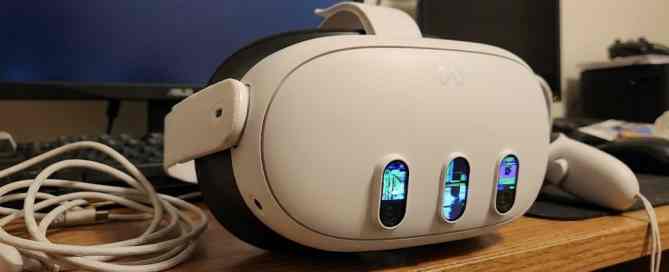 A white virtual reality headset with three front-facing cameras is placed on a wooden desk, with a VR controller and a USB cable nearby. Computer monitors, a keyboard, and other electronic devices are visible in the background. The display reflections are visible in the headset's camera lenses.