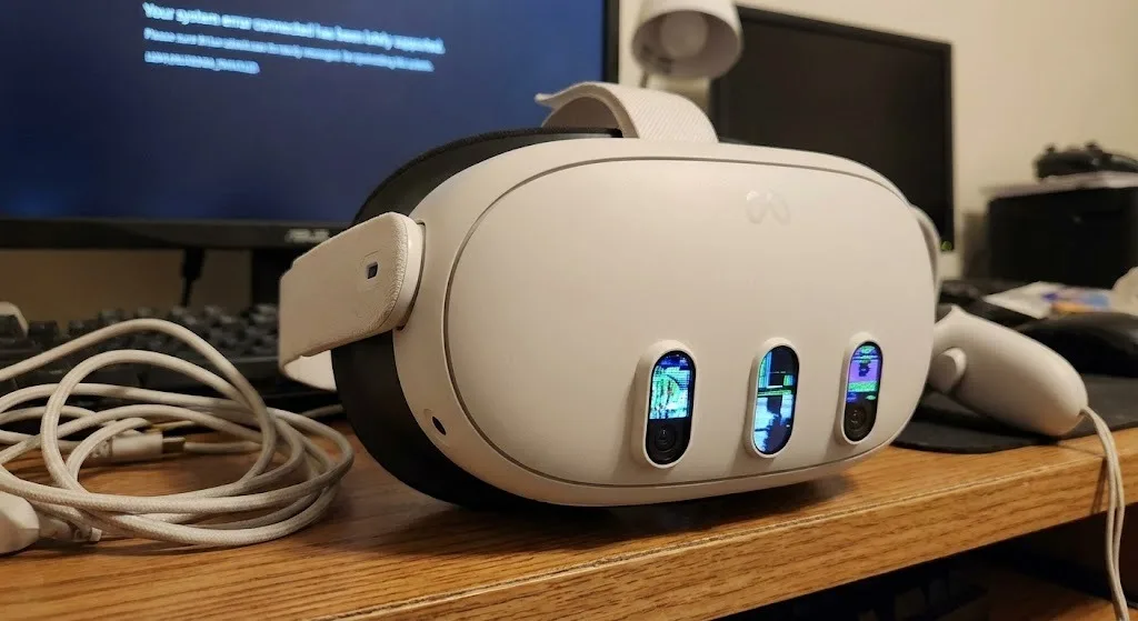 A white virtual reality headset with three front-facing cameras is placed on a wooden desk, with a VR controller and a USB cable nearby. Computer monitors, a keyboard, and other electronic devices are visible in the background. The display reflections are visible in the headset's camera lenses.